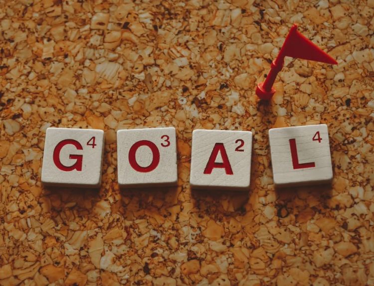 Scrabble tiles forming the word 'goal' with a small red flag on a cork board background.